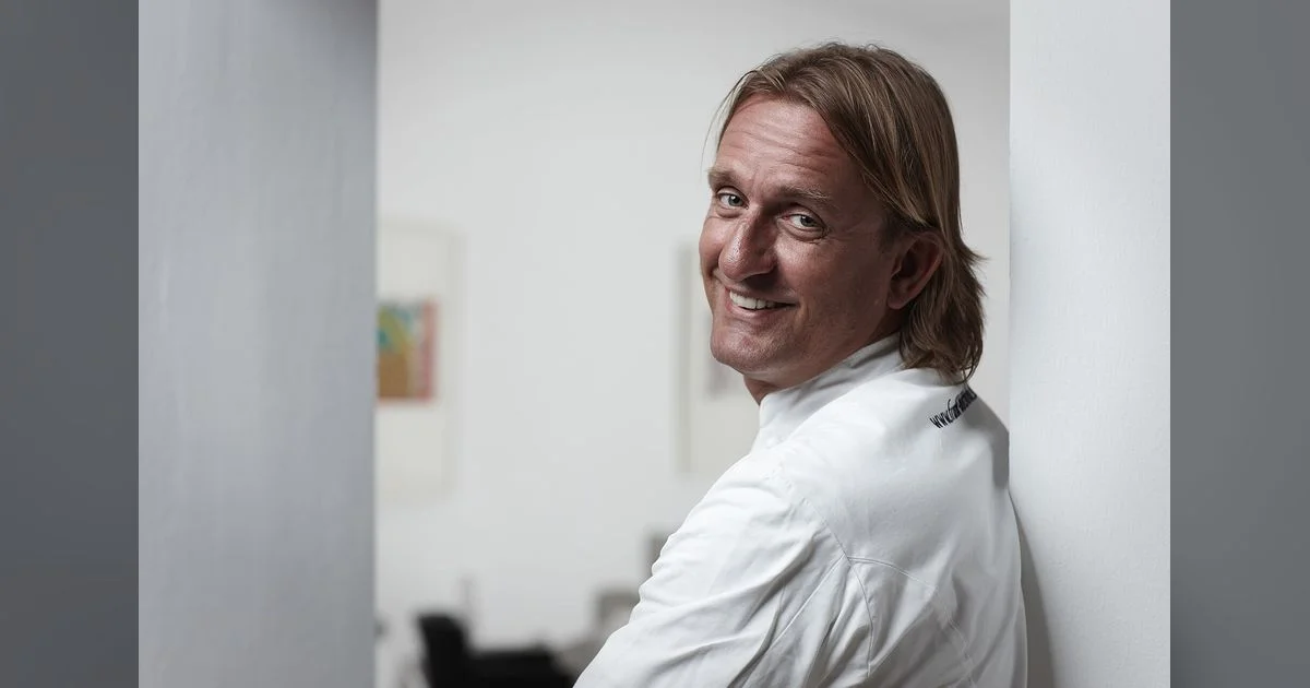 Frank Buchholz in weißer Kochjacke lehnt lächelnd an einer Wand